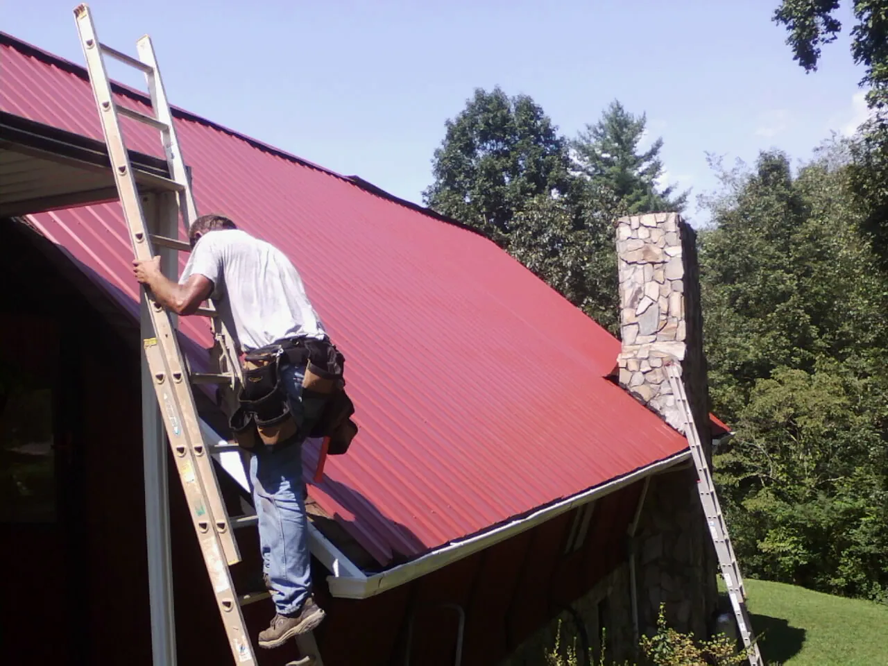 Roofer on a ladder beside a red metal roof and stone chimney — Soffit & Fascia Repair by Roofing Brothers in Morrow